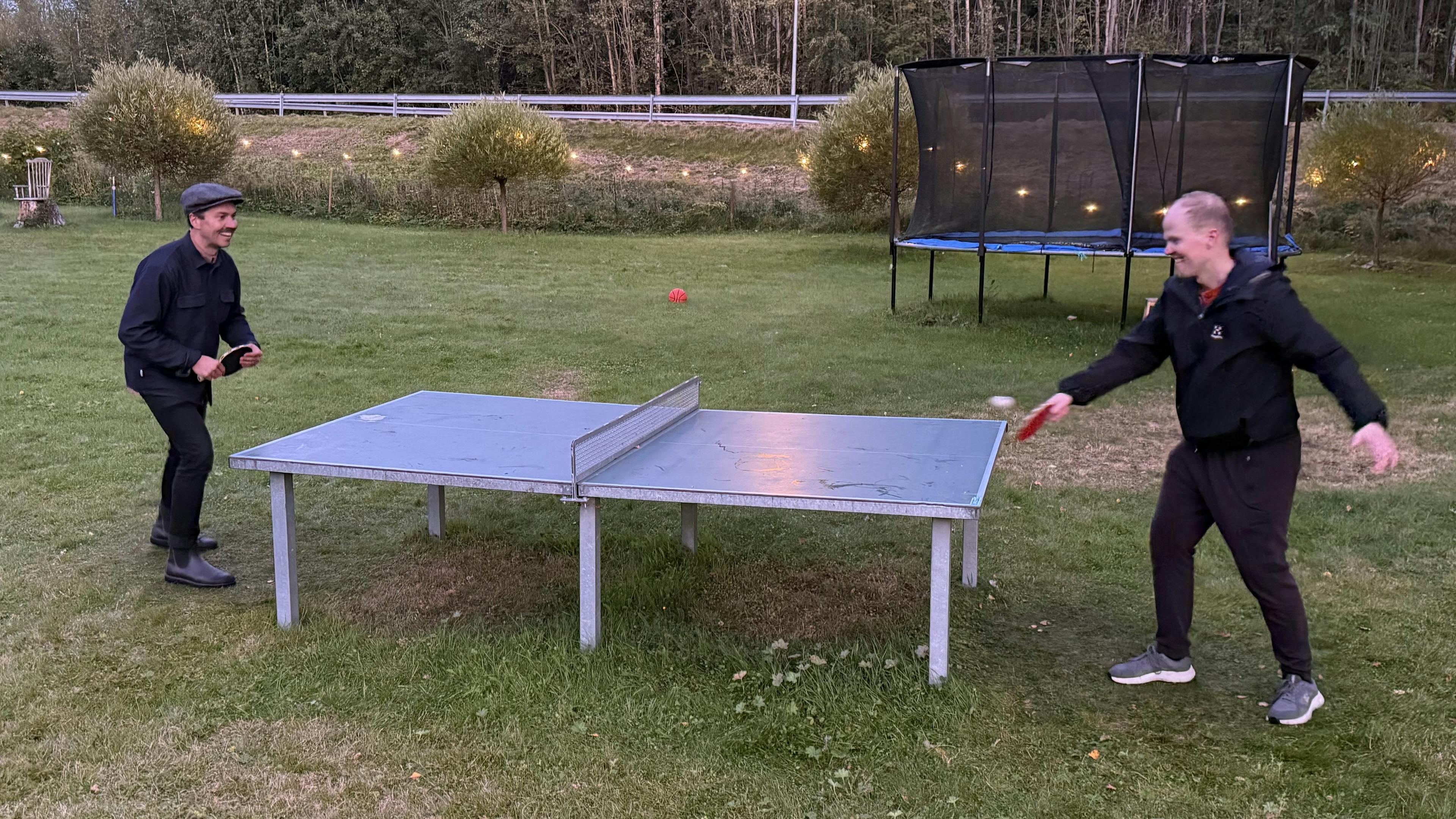 Juho and Hannu playing table tennis outdoors on a grassy lawn at dusk.