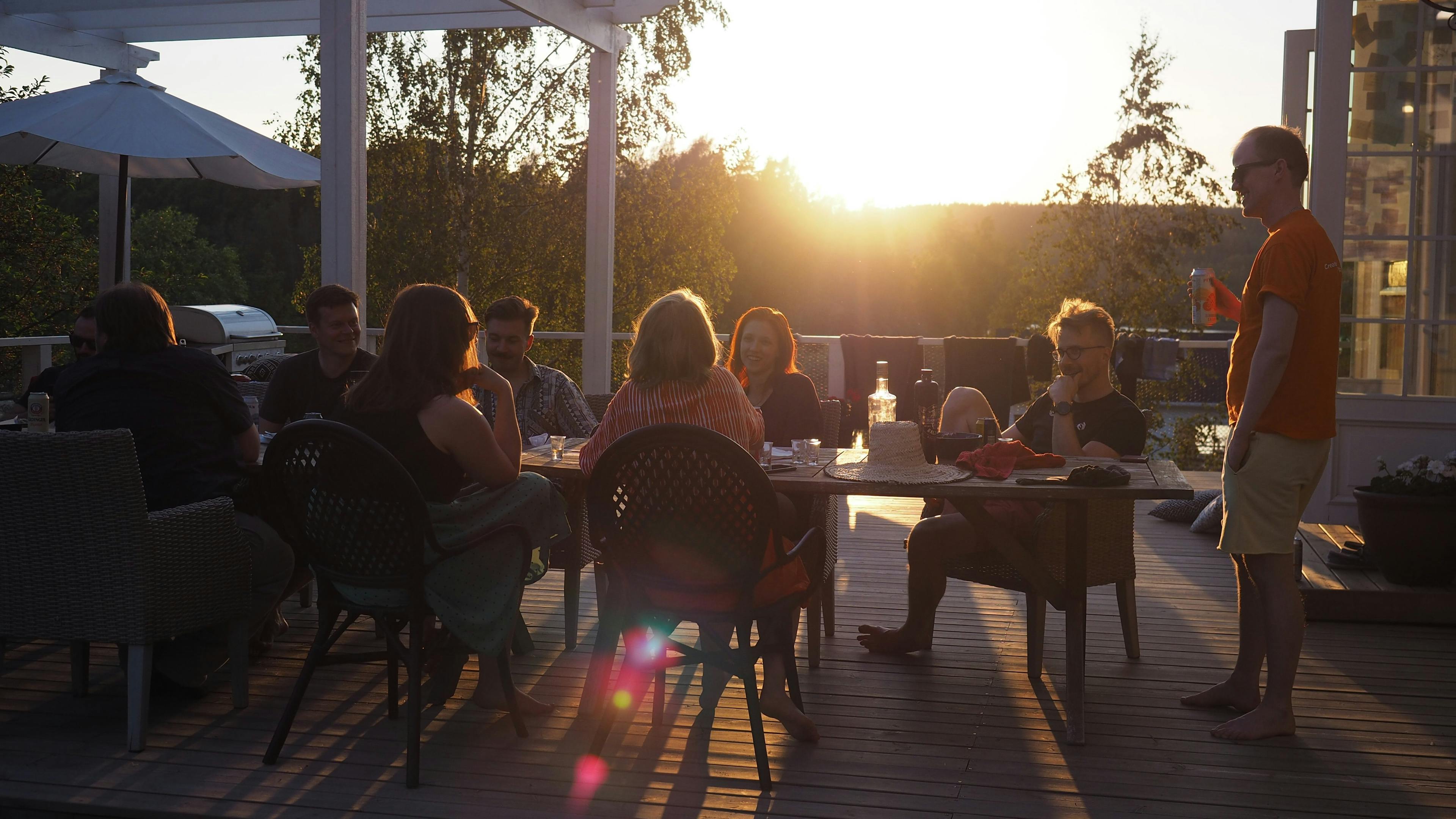 A group of people sitting around a table on a terrace at sunset, talking and enjoying drinks.