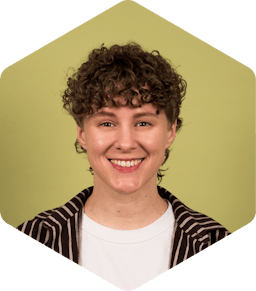 Katri is a white woman with short, curly hair and she's smiling facing the camera. She's wearing a brown, striped Marimekko shirt over a white tee.