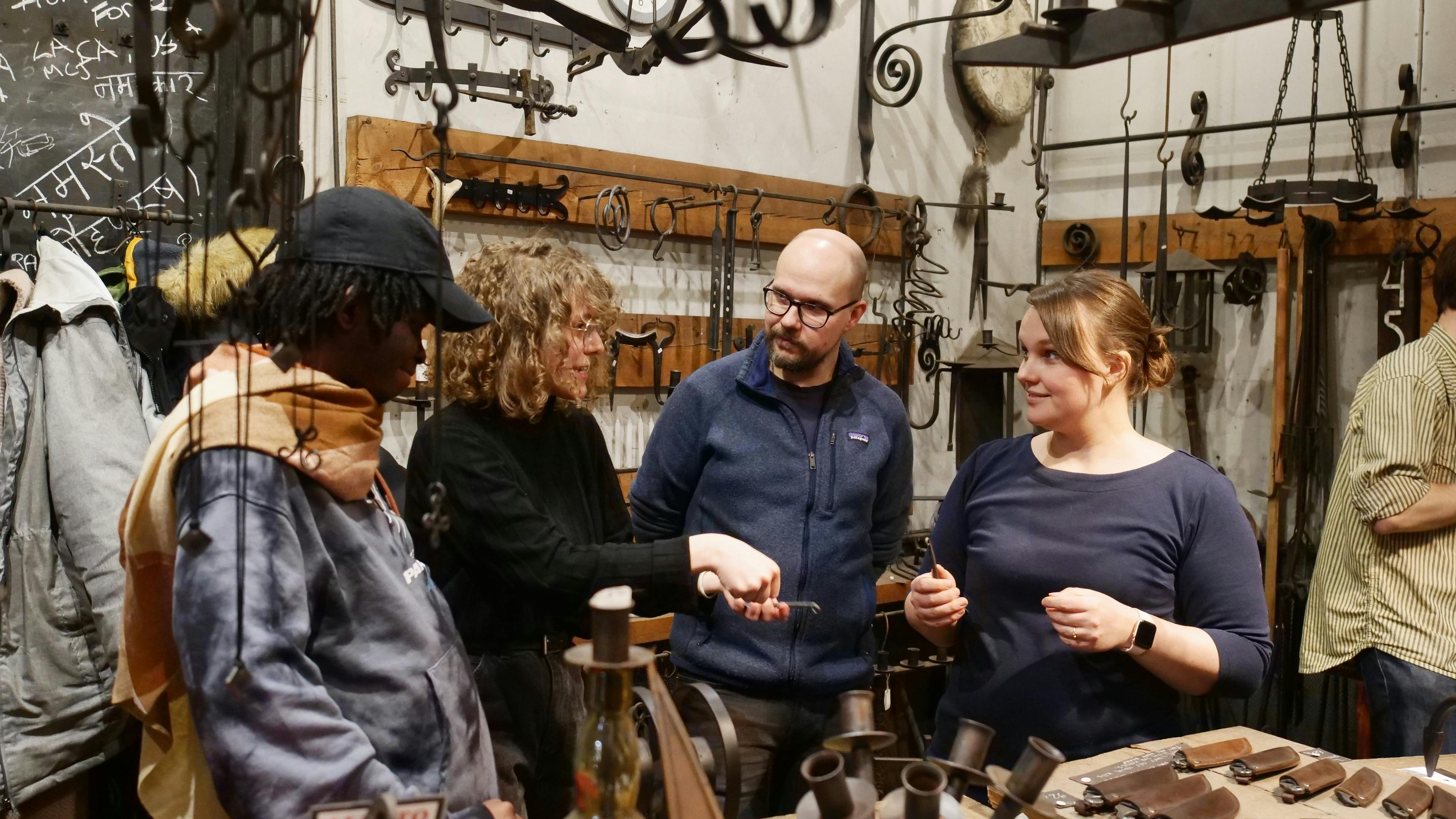 Four people stand together in a blacksmith workshop, talking and holding small hand tools.