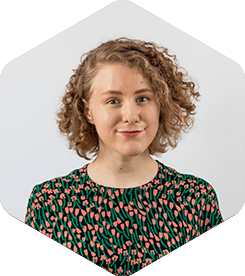 Katri is white woman with curly dark blonde hair. She's wearing a shirt with salmon pink and green flowers.