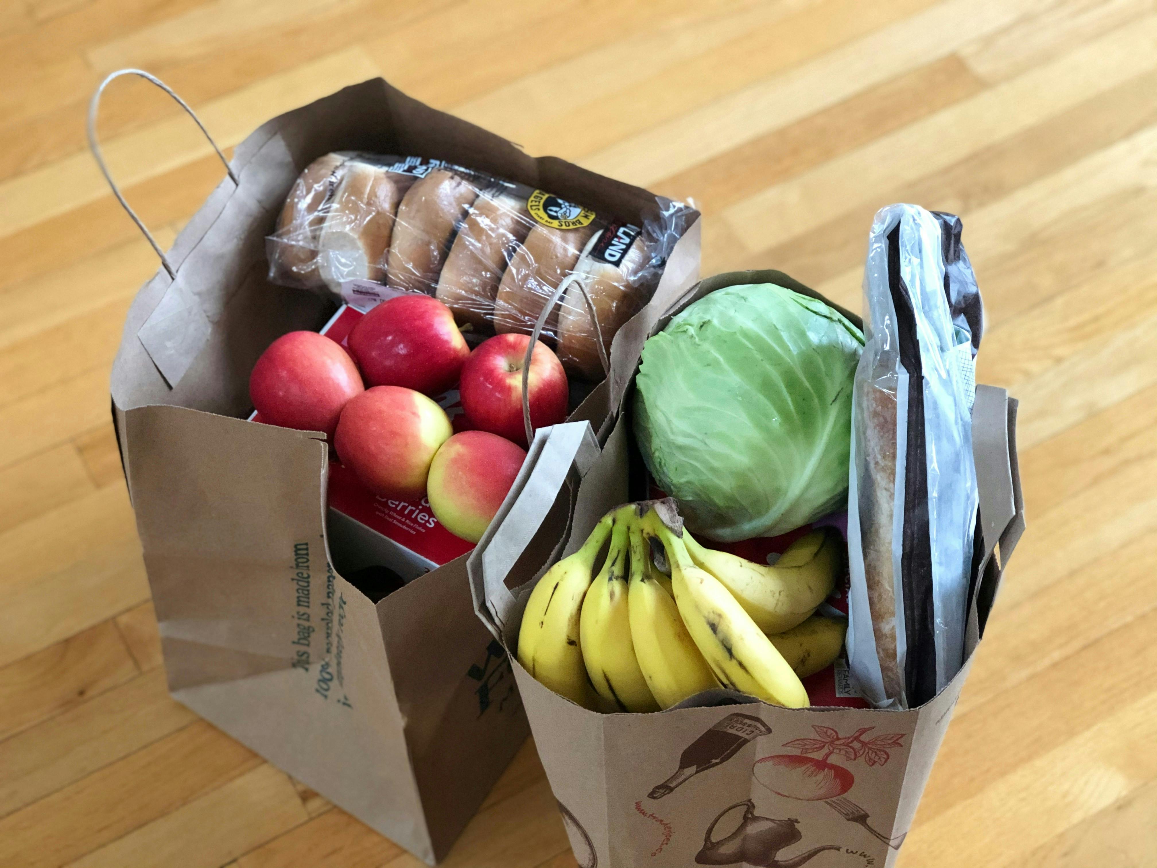 Two paper bags of groceries on a wooden floor. Visible on top are apples, bagels, cabbage, bananas, and a baguette.