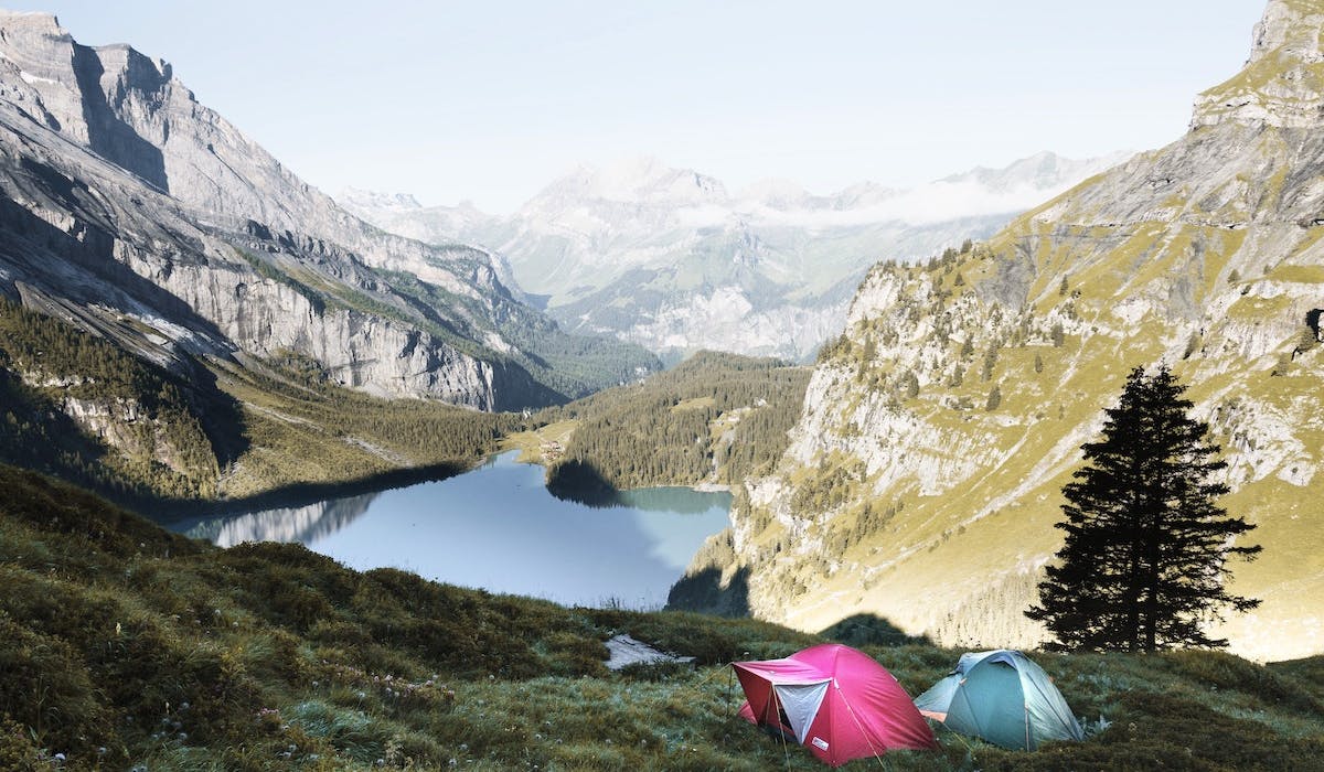 Two tents in a mountain scenery. In between the mountains, beyond the hill the tents are, there's a lake. Photo by Dino Reichmuth on Unsplash.