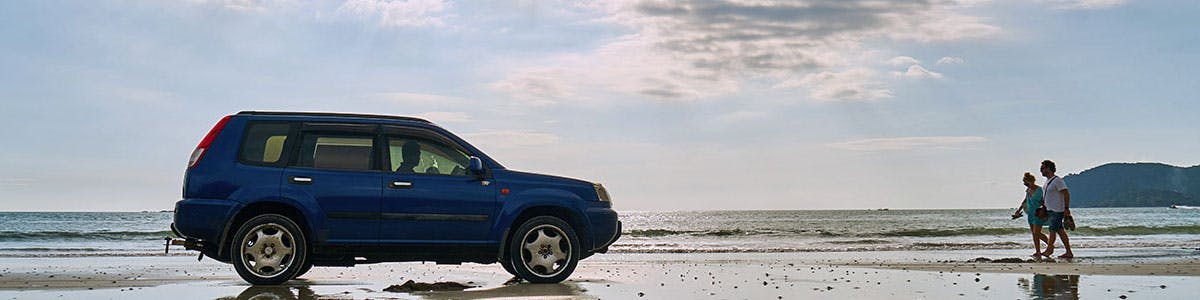 Two people walking from the right towards a blue SUV parked on a beach.
