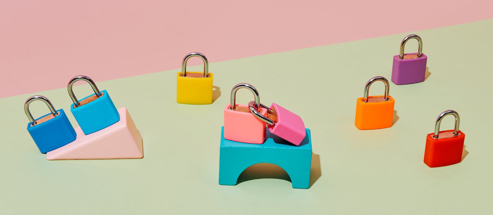 Eight randomly placed locked padlocks of different colors, some of them on top of different building blocks, on a pale green surface, against a pale pink background.