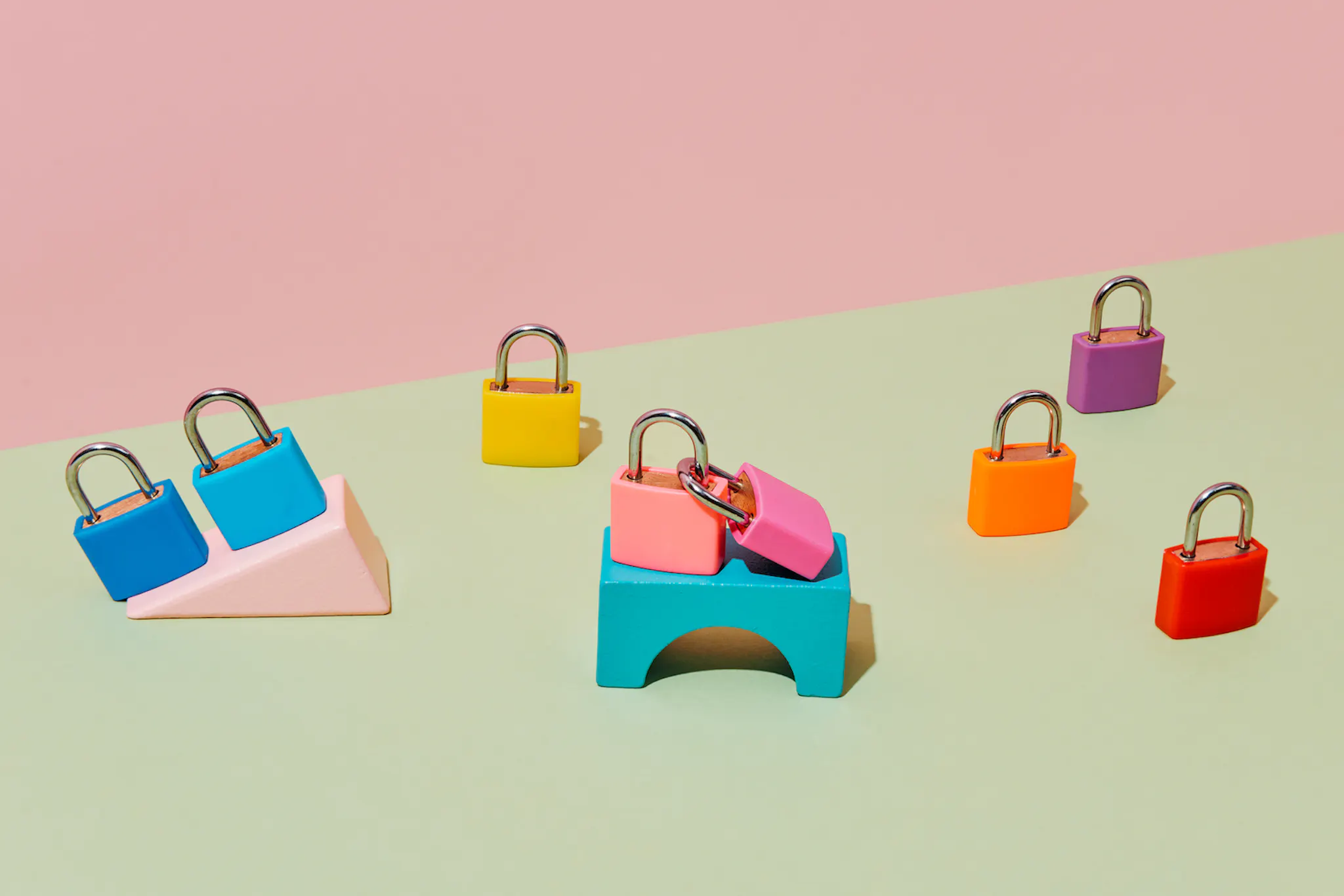 Eight randomly placed locked padlocks of different colors, some of them on top of different building blocks, on a pale green surface, against a pale pink background.