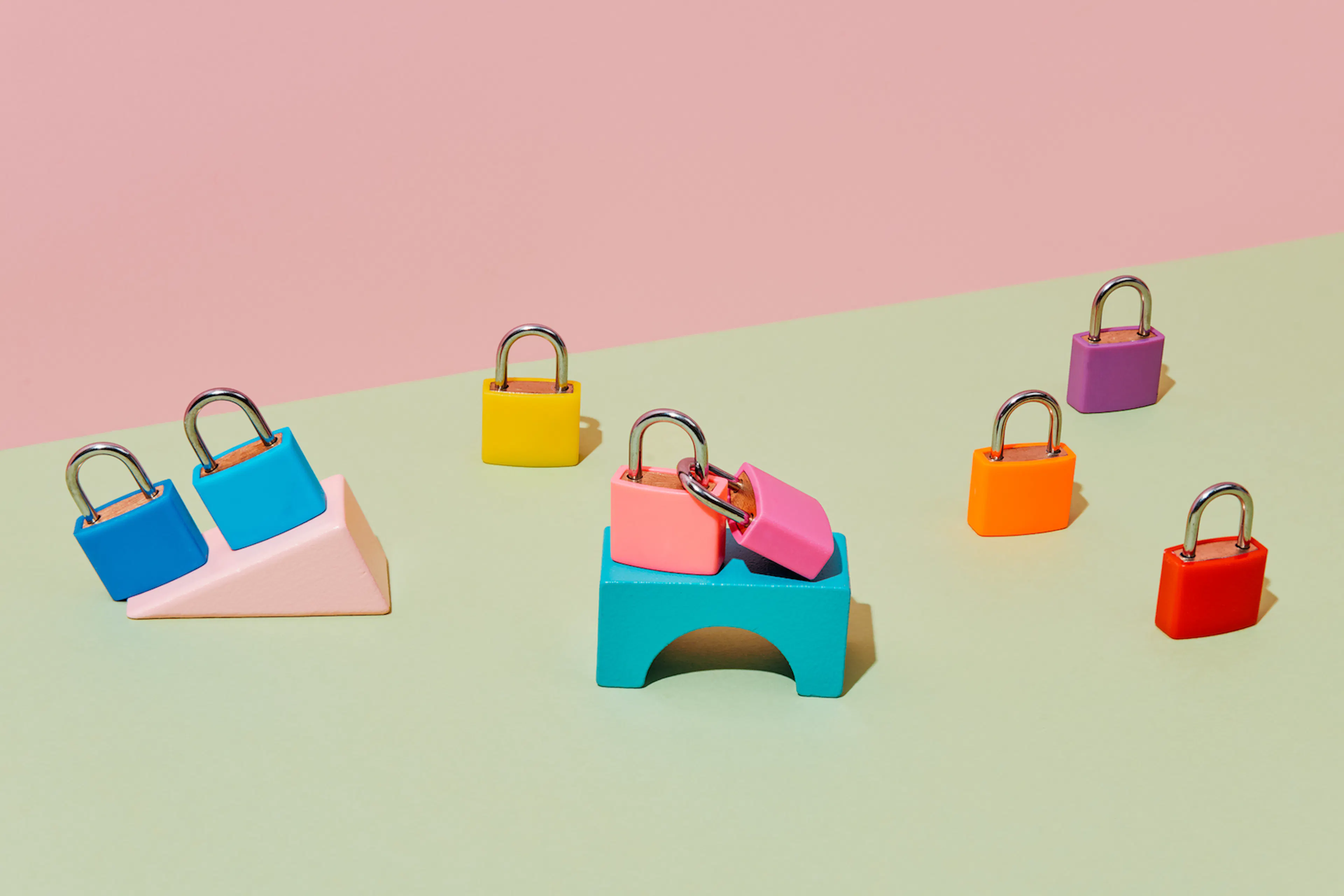 Eight randomly placed locked padlocks of different colors, some of them on top of different building blocks, on a pale green surface, against a pale pink background.