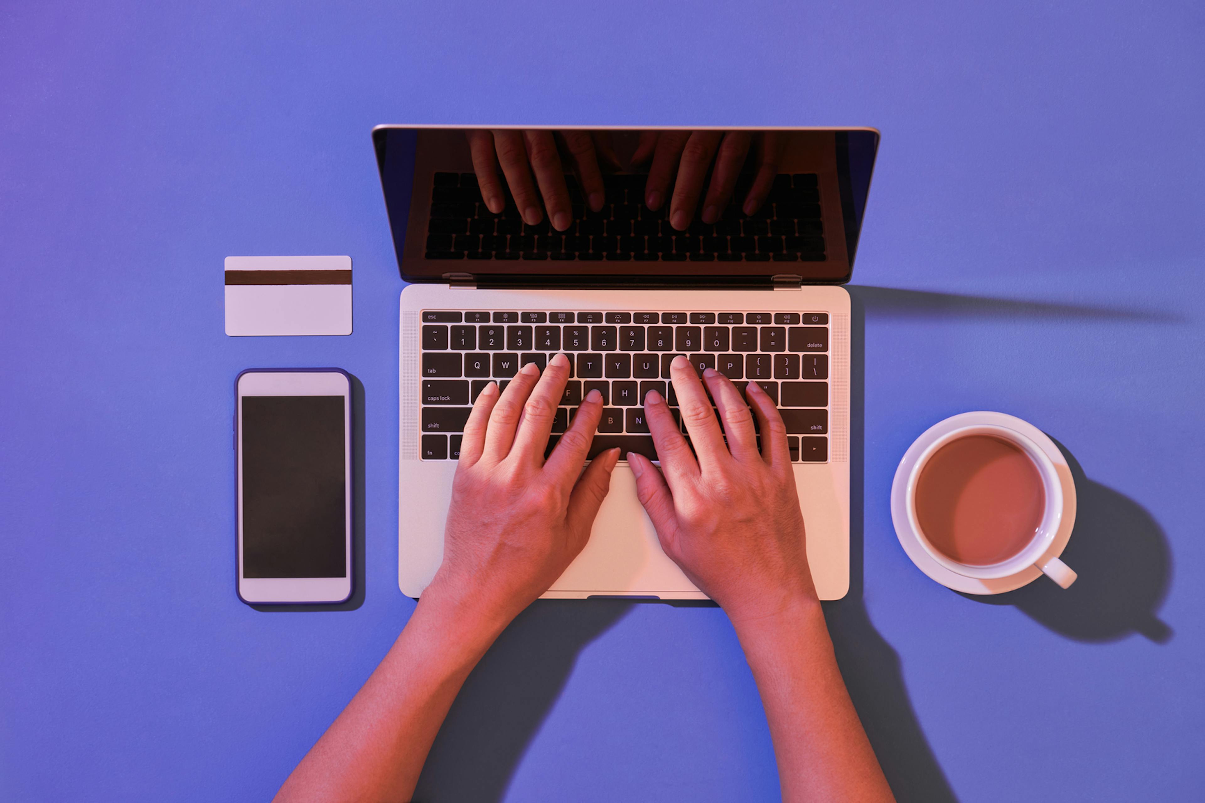 A table top, viewed from above, with an open laptop. A person's hands are on the keys. Beside the laptop are a credit card, a smartphone, and a coffee cup that's half full of coffee milk. Or possibly black tea.