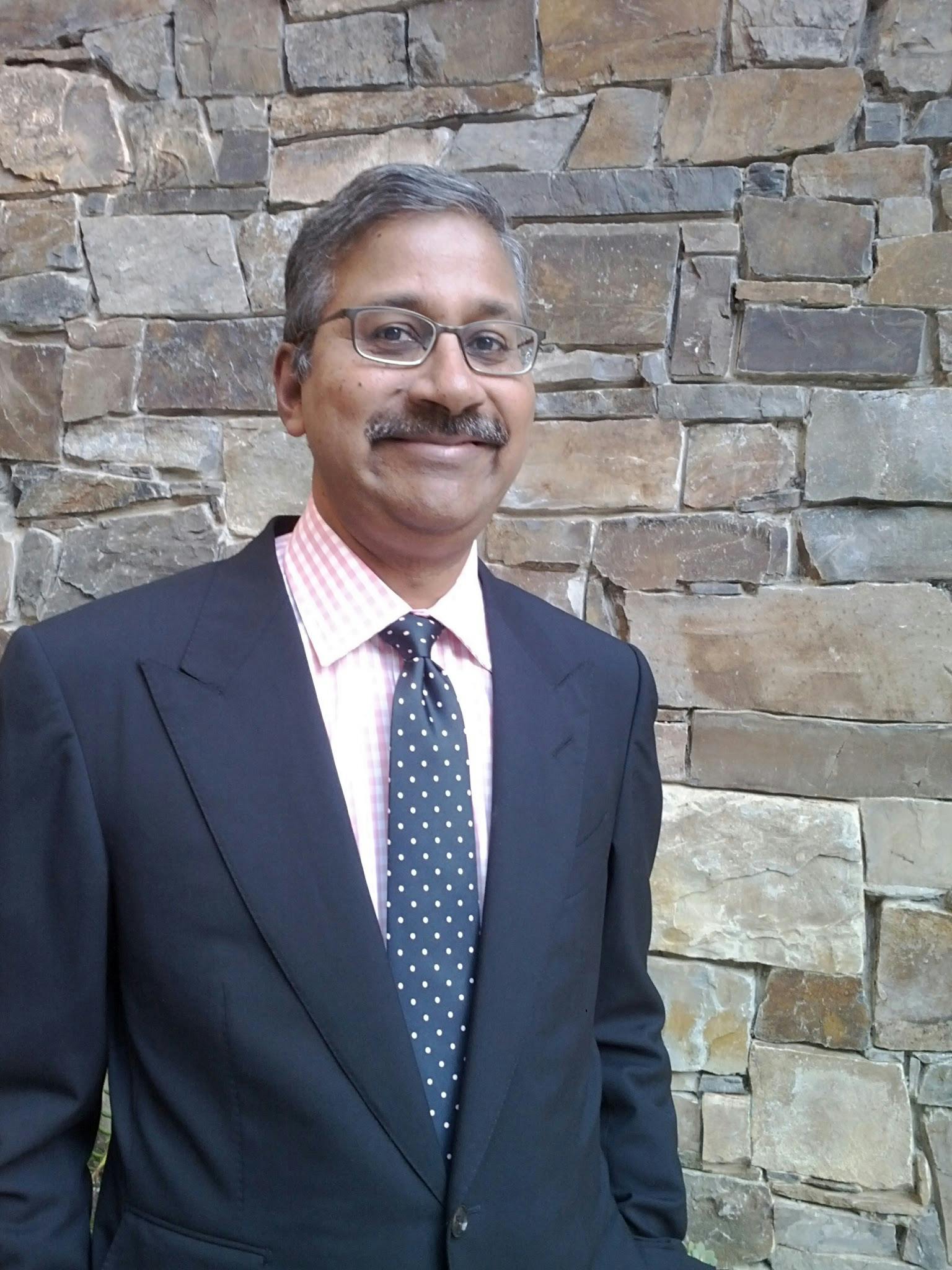 Ram Shriram is pictured standing in front of a brick wall. He is wearing a suit and tie and is smiling.