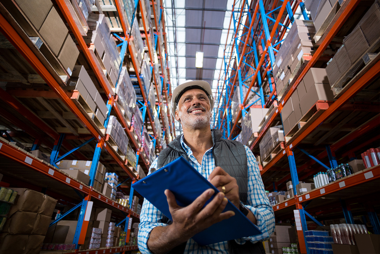 Man wearing a hard hat carrying a blue clipboard walking between two warehouse aisles 