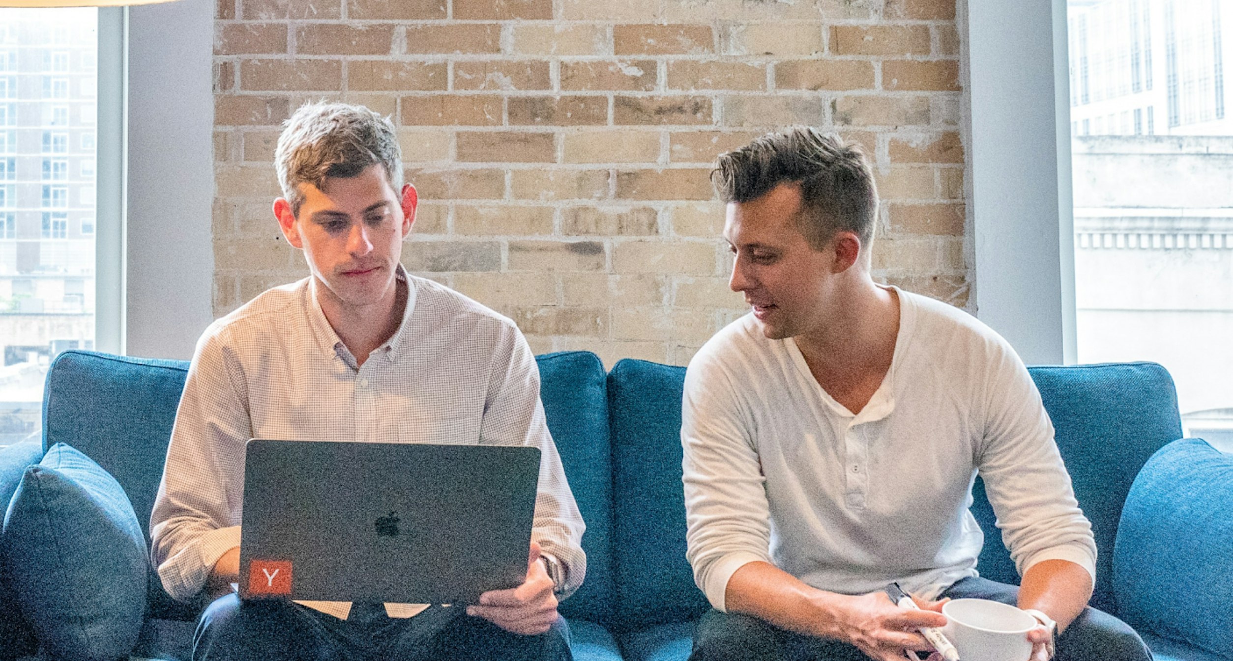 Two men sitting on a sofa, working on a laptop computer on the left-hand man's lap