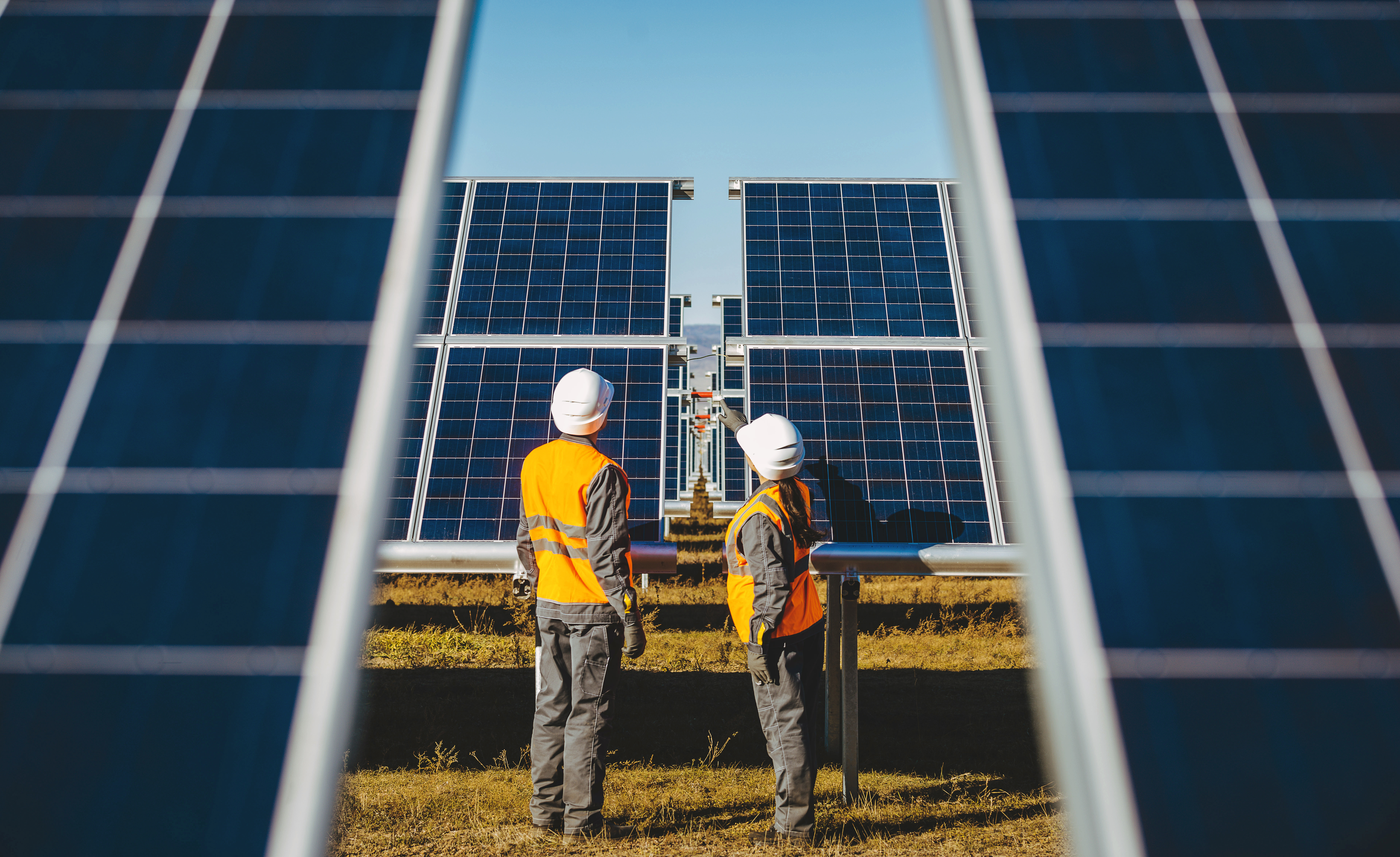 2 people facing away from the camera and dressed in work clothes, talking in front of solar panels on a solar farm