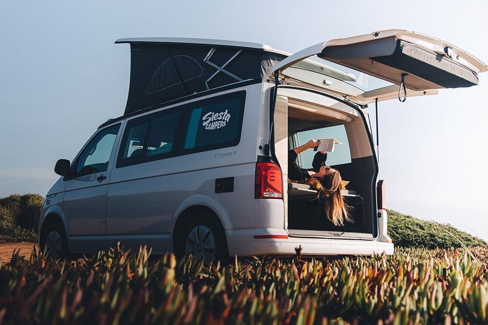 Uma mulher lê um livro na parte de trás da sua campervan, desfrutando do ar fresco enquanto relaxa no seu ambiente sem tecnologia.