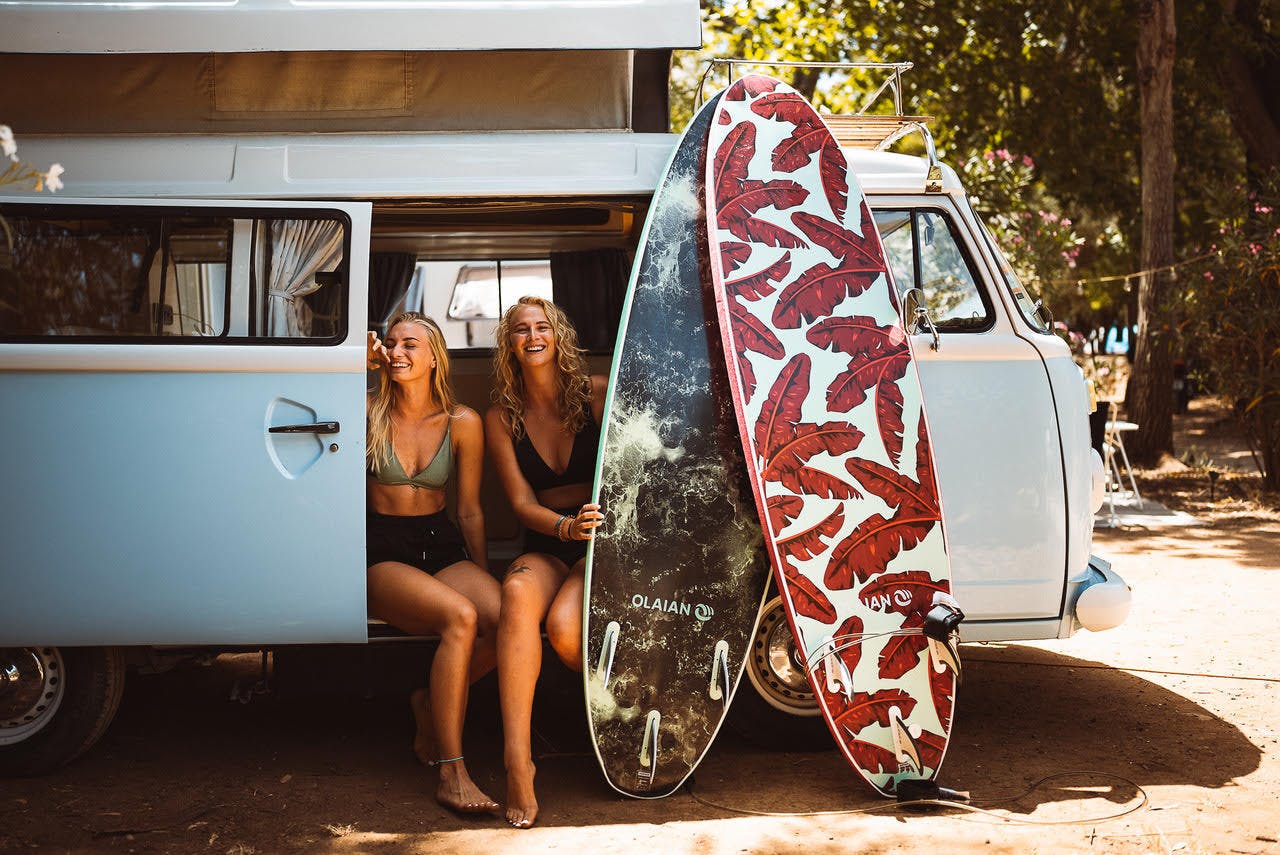 Zwei Surfer/innen posieren für ein Erinnerungsfoto vor ihrem alten VW-Campervan, bevor sie sich für ihre morgendliche Surf-Session bereit machen.