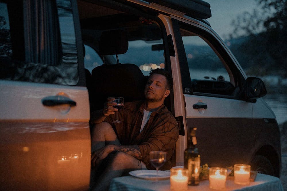 Un homme se détend à l’arrière de son camping-car avec un verre de vin après une journée d’activités en plein air, dans une ambiance chaleureuse, à la lumière de bougies.