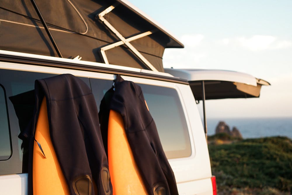 Deux combinaisons de plongée sèchent sur deux planches de surf orange posées contre un camping-car Siesta Campers blanc garé près de l'océan.