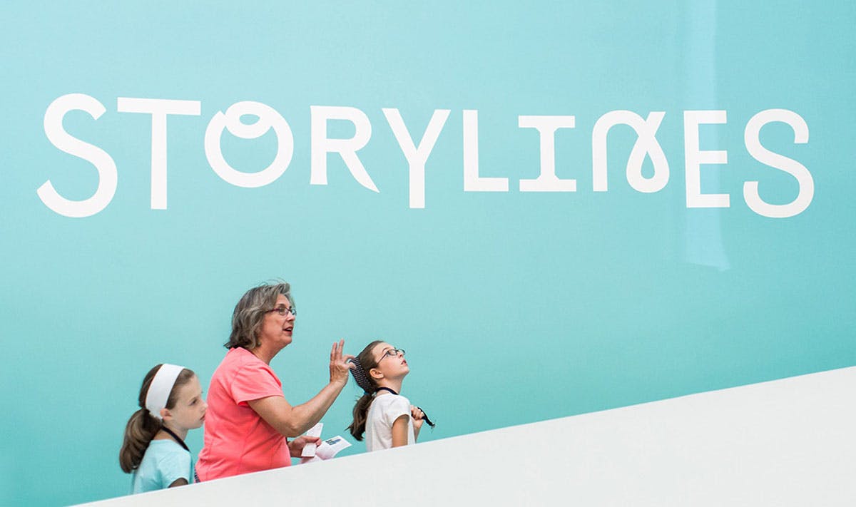 Woman leading two young girls up stairs next to blue wall that reads "Storylines"