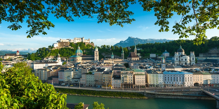 Panoramablick auf eine historische Altstadt mit Festung auf einem Hügel über einem Fluss. Im Vordergrund rahmen grüne Baumäste die Szene, während sich Kirchenkuppeln und Türme vor einer bergigen Kulisse erheben.