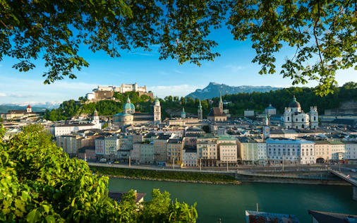 Panoramablick auf eine historische Altstadt mit Festung auf einem Hügel über einem Fluss. Im Vordergrund rahmen grüne Baumäste die Szene, während sich Kirchenkuppeln und Türme vor einer bergigen Kulisse erheben.