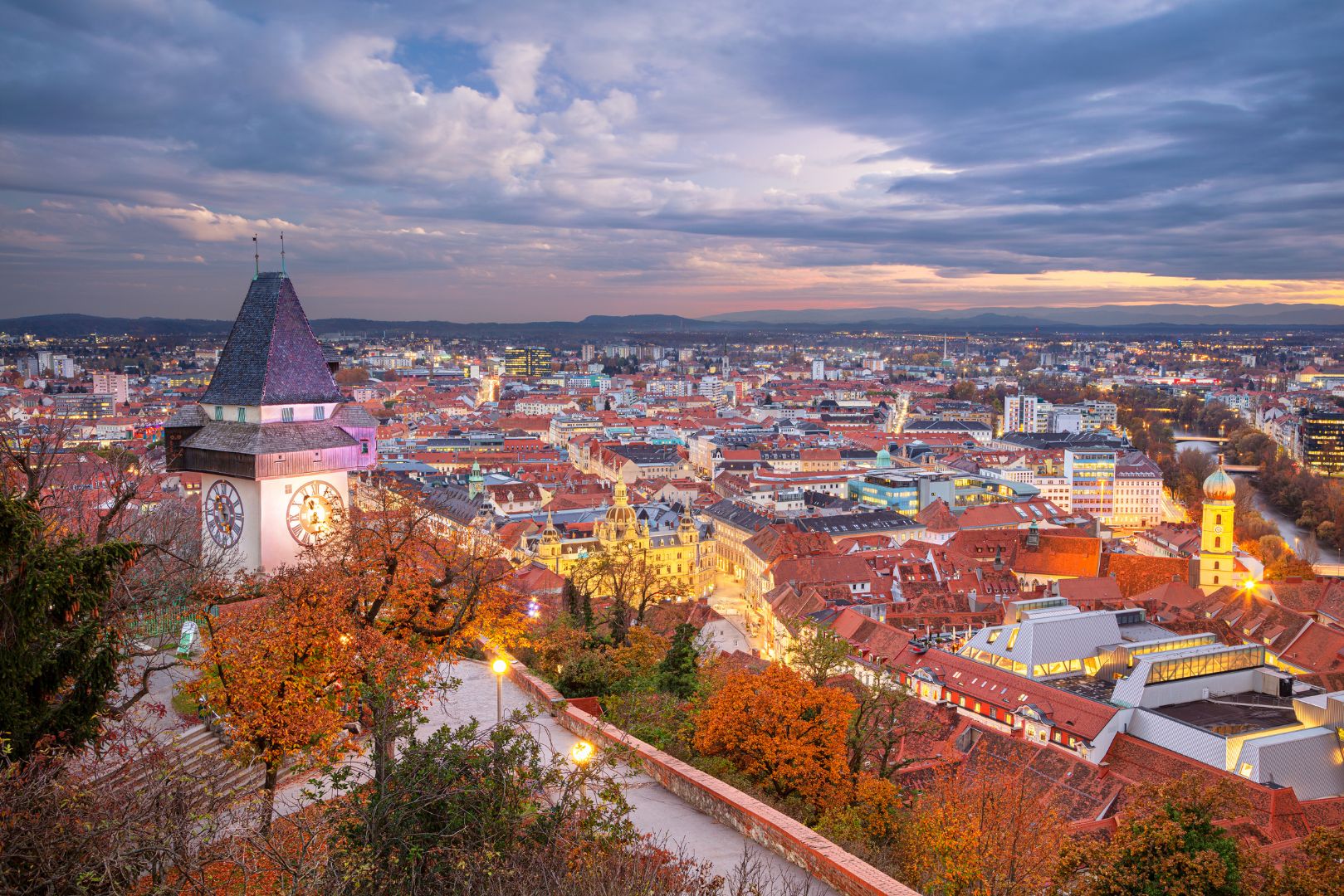 Panoramablick über eine historische Altstadt mit roten Ziegeldächern und Kirchtürmen. Im Vordergrund rahmen eine Steinmauer und herbstlich gefärbte Bäume die Szene, während eine markante Kirche mit steilem Dach über der Stadt emporragt. In der Ferne öffnet sich die Landschaft zu einer weiten Ebene unter einem sanft beleuchteten Abendhimmel.