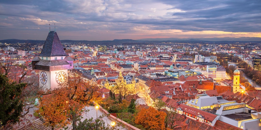 Panoramablick über eine historische Altstadt mit roten Ziegeldächern und Kirchtürmen. Im Vordergrund rahmen eine Steinmauer und herbstlich gefärbte Bäume die Szene, während eine markante Kirche mit steilem Dach über der Stadt emporragt. In der Ferne öffnet sich die Landschaft zu einer weiten Ebene unter einem sanft beleuchteten Abendhimmel.