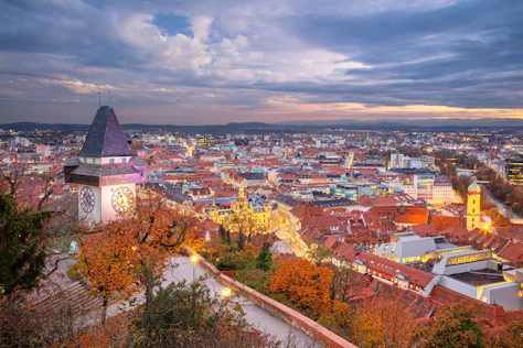 Panoramablick über eine historische Altstadt mit roten Ziegeldächern und Kirchtürmen. Im Vordergrund rahmen eine Steinmauer und herbstlich gefärbte Bäume die Szene, während eine markante Kirche mit steilem Dach über der Stadt emporragt. In der Ferne öffnet sich die Landschaft zu einer weiten Ebene unter einem sanft beleuchteten Abendhimmel.