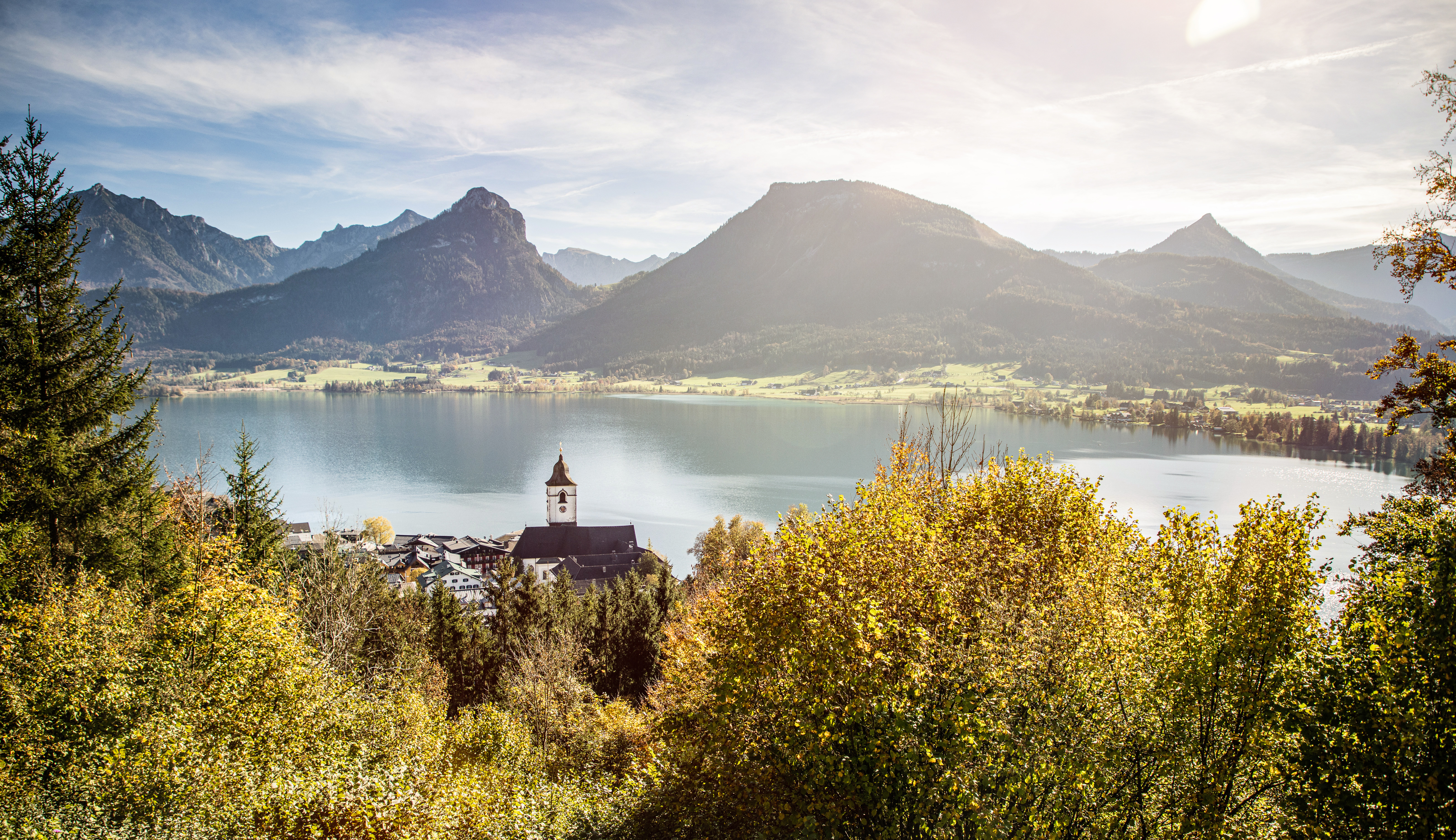 View from a wooded hillside over a clear lake, with gentle meadows and striking mountain peaks in the background. In the foreground lies a small village with a church featuring an onion dome, surrounded by golden autumn trees in warm sunlight.