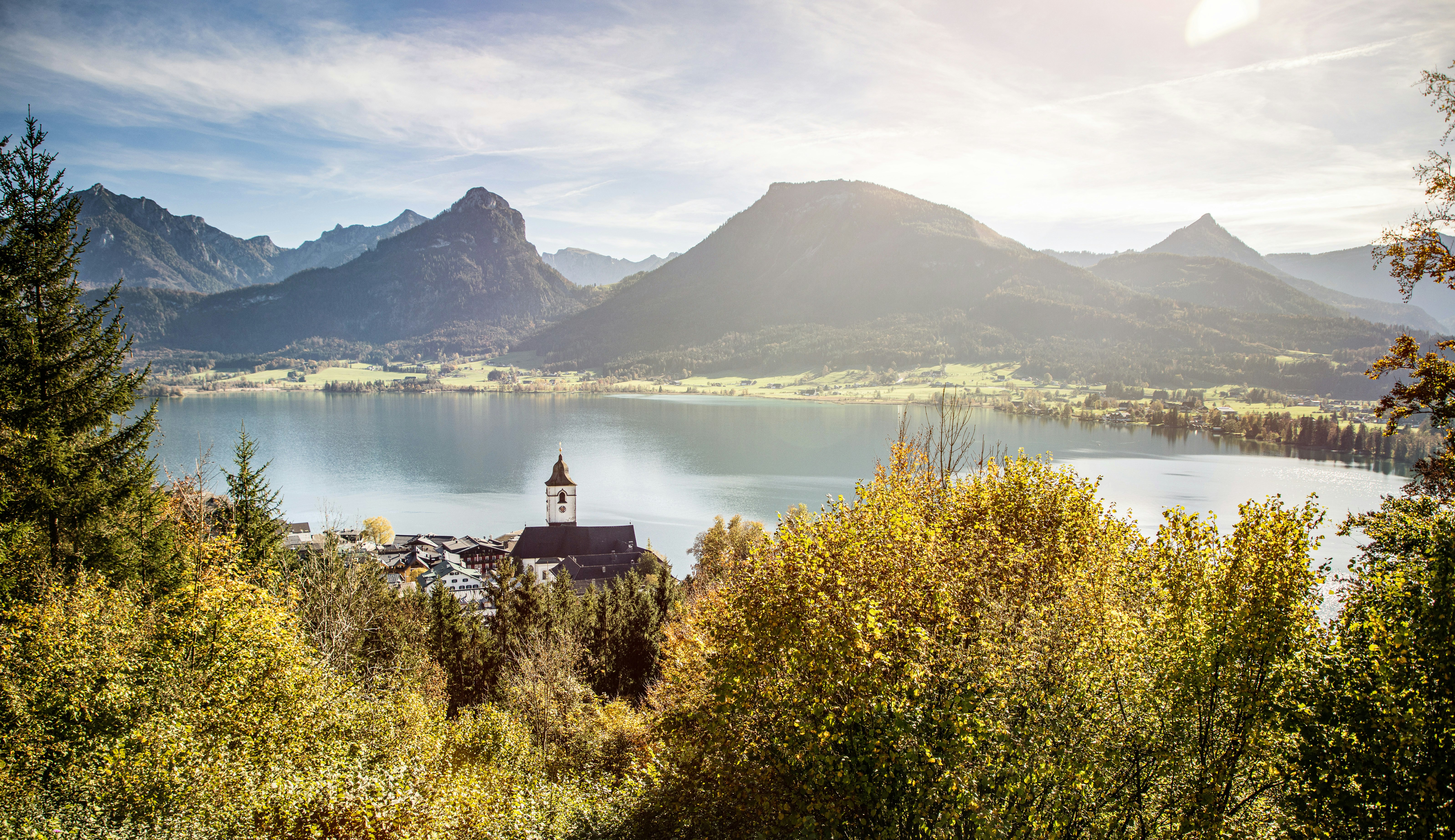 Blick von einem bewaldeten Hang auf einen klaren See, dahinter sanfte Wiesen und markante Berggipfel. Im Vordergrund liegt ein kleines Dorf mit einer Kirche und Zwiebelturm, umgeben von goldgelben Herbstbäumen im warmen Sonnenlicht.