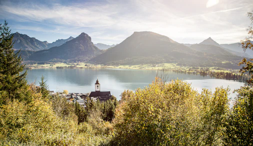 Blick von einem bewaldeten Hang auf einen klaren See, dahinter sanfte Wiesen und markante Berggipfel. Im Vordergrund liegt ein kleines Dorf mit einer Kirche und Zwiebelturm, umgeben von goldgelben Herbstbäumen im warmen Sonnenlicht.