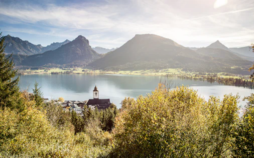 Blick von einem bewaldeten Hang auf einen klaren See, dahinter sanfte Wiesen und markante Berggipfel. Im Vordergrund liegt ein kleines Dorf mit einer Kirche und Zwiebelturm, umgeben von goldgelben Herbstbäumen im warmen Sonnenlicht.