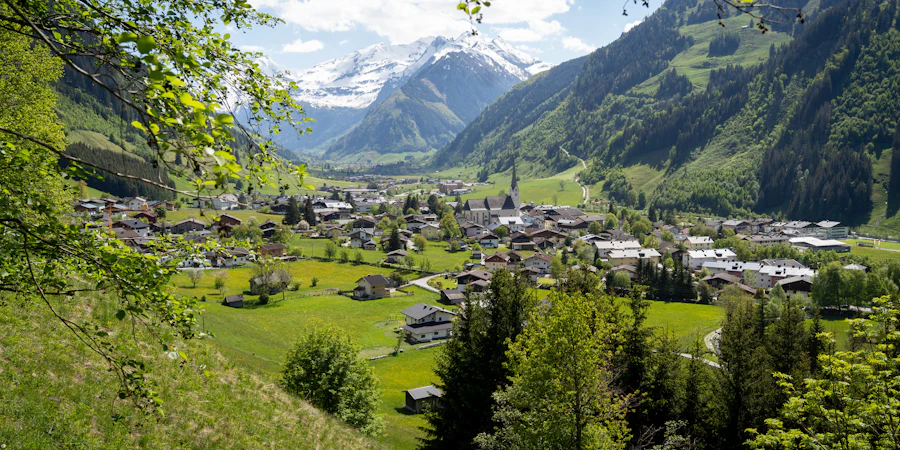 Erhöhter Blick auf ein alpines Dorf in einem grünen Tal, umgeben von bewaldeten Hängen und schneebedeckten Bergen im Hintergrund. Traditionelle Häuser und eine Kirche mit hohem Turm liegen eingebettet in weite Wiesen unter einem klaren blauen Himmel.