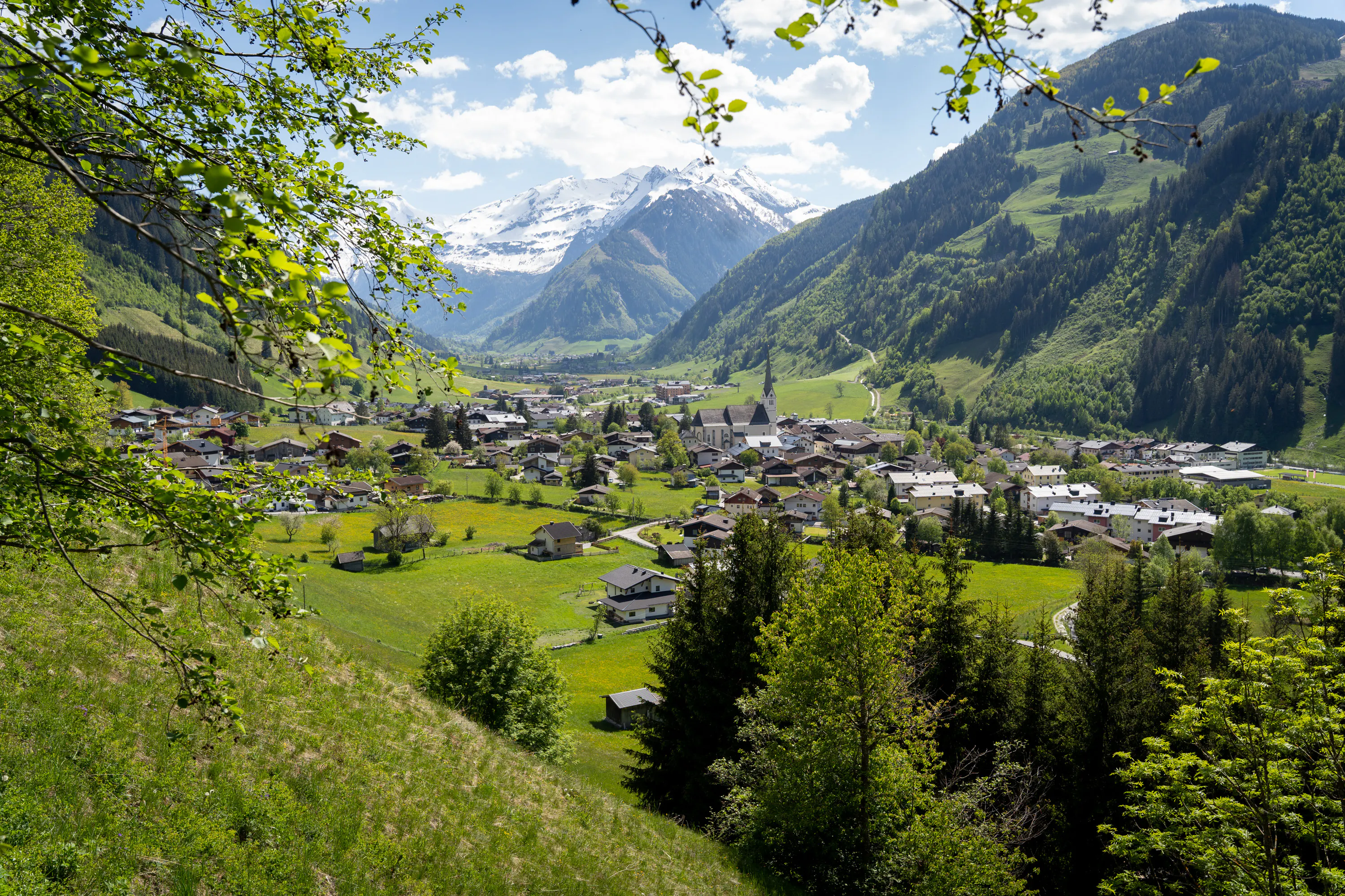 Elevated view of an alpine village nestled in a green valley, surrounded by forested hills and snow-capped mountains in the distance. Traditional houses and a church with a tall steeple sit among open meadows under a bright blue sky.