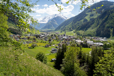 Erhöhter Blick auf ein alpines Dorf in einem grünen Tal, umgeben von bewaldeten Hängen und schneebedeckten Bergen im Hintergrund. Traditionelle Häuser und eine Kirche mit hohem Turm liegen eingebettet in weite Wiesen unter einem klaren blauen Himmel.