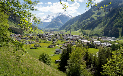 Erhöhter Blick auf ein alpines Dorf in einem grünen Tal, umgeben von bewaldeten Hängen und schneebedeckten Bergen im Hintergrund. Traditionelle Häuser und eine Kirche mit hohem Turm liegen eingebettet in weite Wiesen unter einem klaren blauen Himmel.