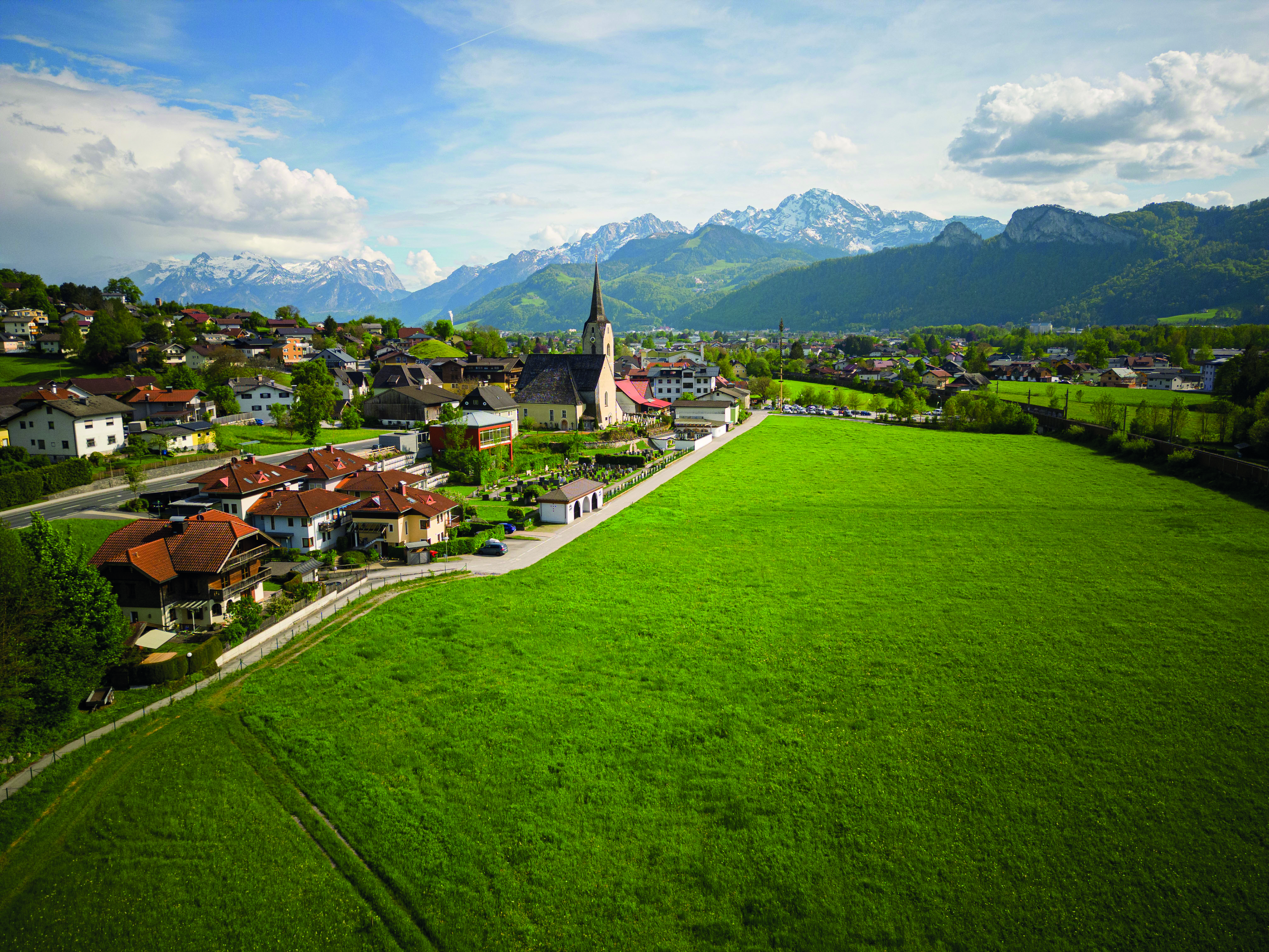 Elevated view of a rural village with a church and prominent steeple, surrounded by wide green fields. Forested hills and snow-capped mountains rise in the background beneath a partly cloudy sky.