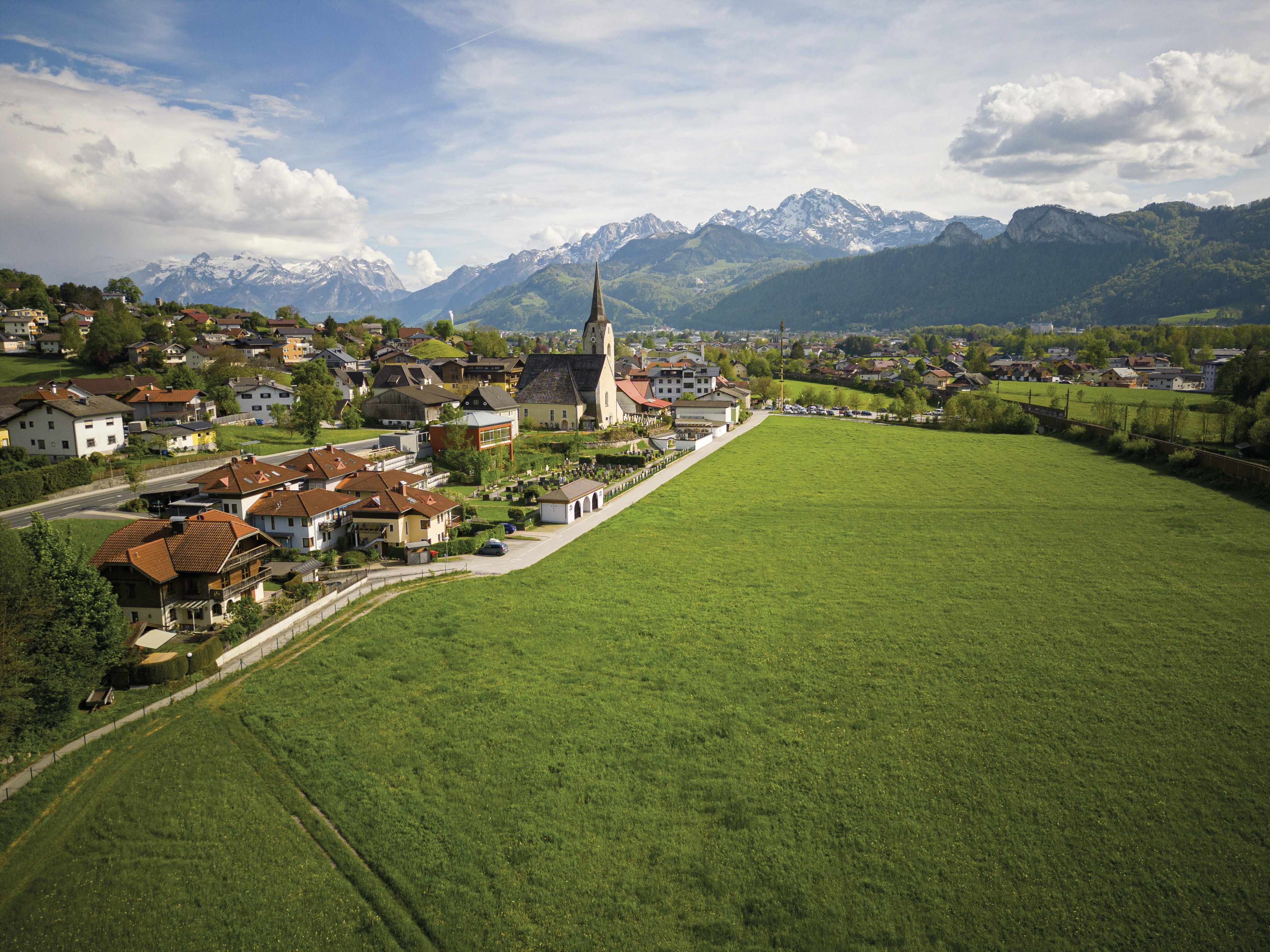 Erhöhter Blick auf eine ländliche Ortschaft mit Kirche und markantem Kirchturm, umgeben von weitläufigen grünen Wiesen. Im Hintergrund erheben sich bewaldete Hügel und schneebedeckte Berge unter einem teilweise bewölkten Himmel.