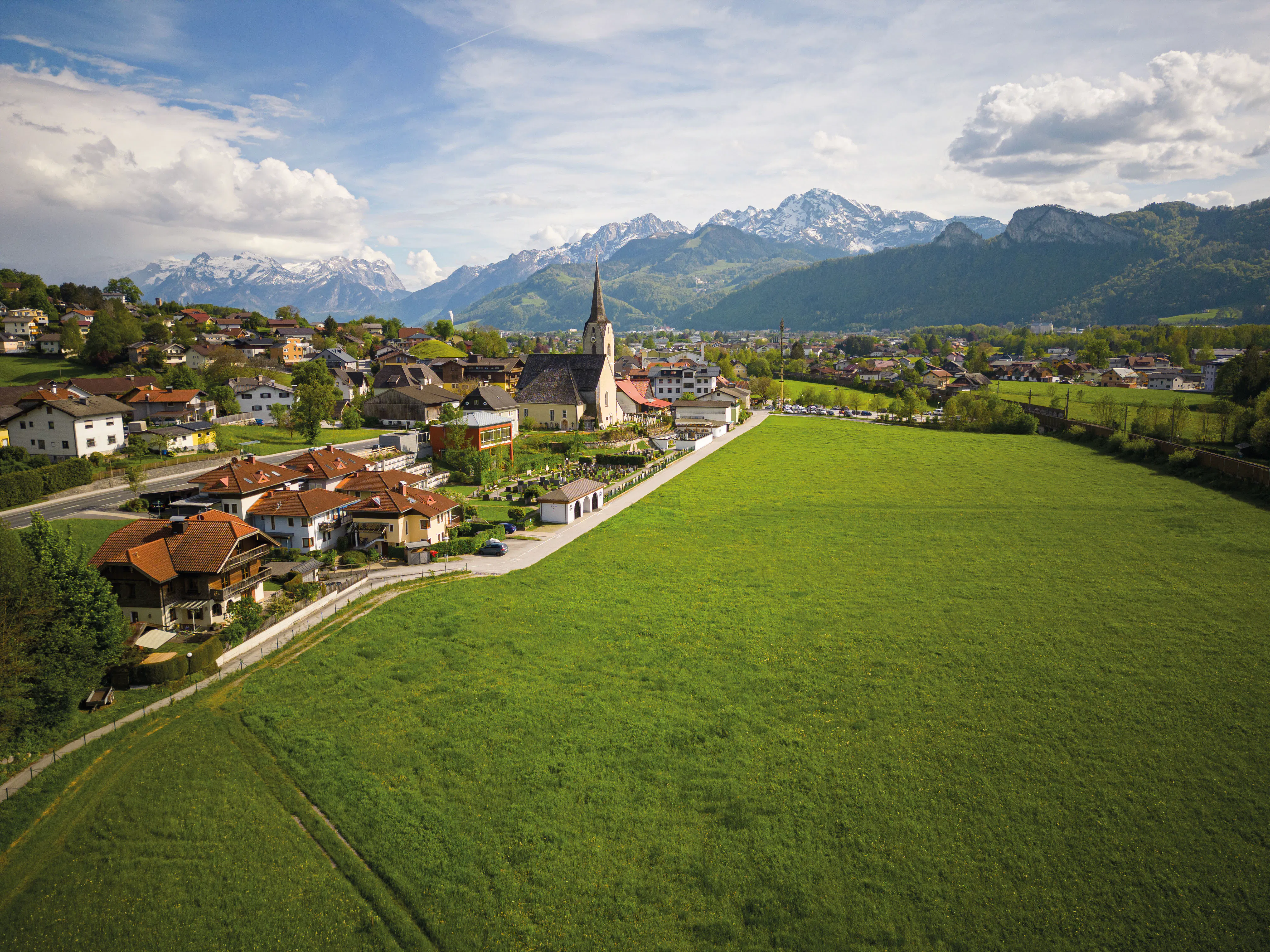 Elevated view of a rural village with a church and prominent steeple, surrounded by wide green fields. Forested hills and snow-capped mountains rise in the background beneath a partly cloudy sky.