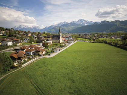 Erhöhter Blick auf eine ländliche Ortschaft mit Kirche und markantem Kirchturm, umgeben von weitläufigen grünen Wiesen. Im Hintergrund erheben sich bewaldete Hügel und schneebedeckte Berge unter einem teilweise bewölkten Himmel.