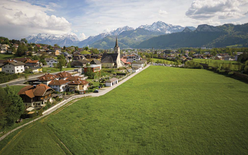 Erhöhter Blick auf eine ländliche Ortschaft mit Kirche und markantem Kirchturm, umgeben von weitläufigen grünen Wiesen. Im Hintergrund erheben sich bewaldete Hügel und schneebedeckte Berge unter einem teilweise bewölkten Himmel.