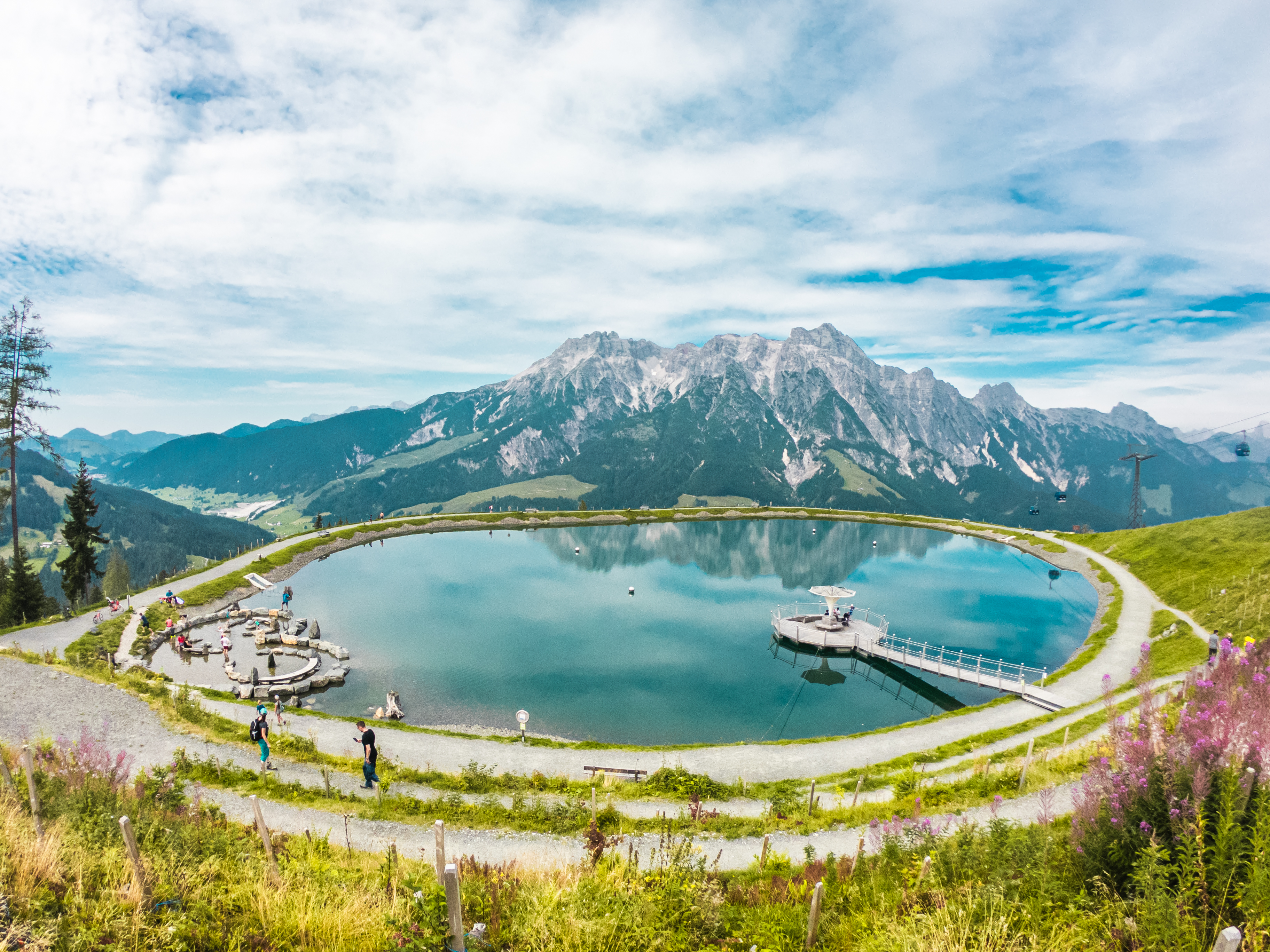 Panoramic view of a circular mountain reservoir surrounded by walking paths and green alpine meadows. Visitors stroll along the shore, while dramatic rocky peaks rise in the background under a partly cloudy sky.