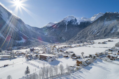 Panoramablick auf ein verschneites Bergdorf mit Kirche und verstreuten Häusern in einem weiten Tal. Umgeben von schneebedeckten Wäldern und hohen Gipfeln liegt die Ortschaft unter strahlendem Sonnenschein und klarem blauem Himmel.
