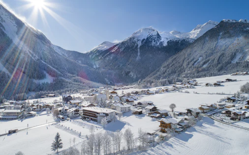 Panoramablick auf ein verschneites Bergdorf mit Kirche und verstreuten Häusern in einem weiten Tal. Umgeben von schneebedeckten Wäldern und hohen Gipfeln liegt die Ortschaft unter strahlendem Sonnenschein und klarem blauem Himmel.