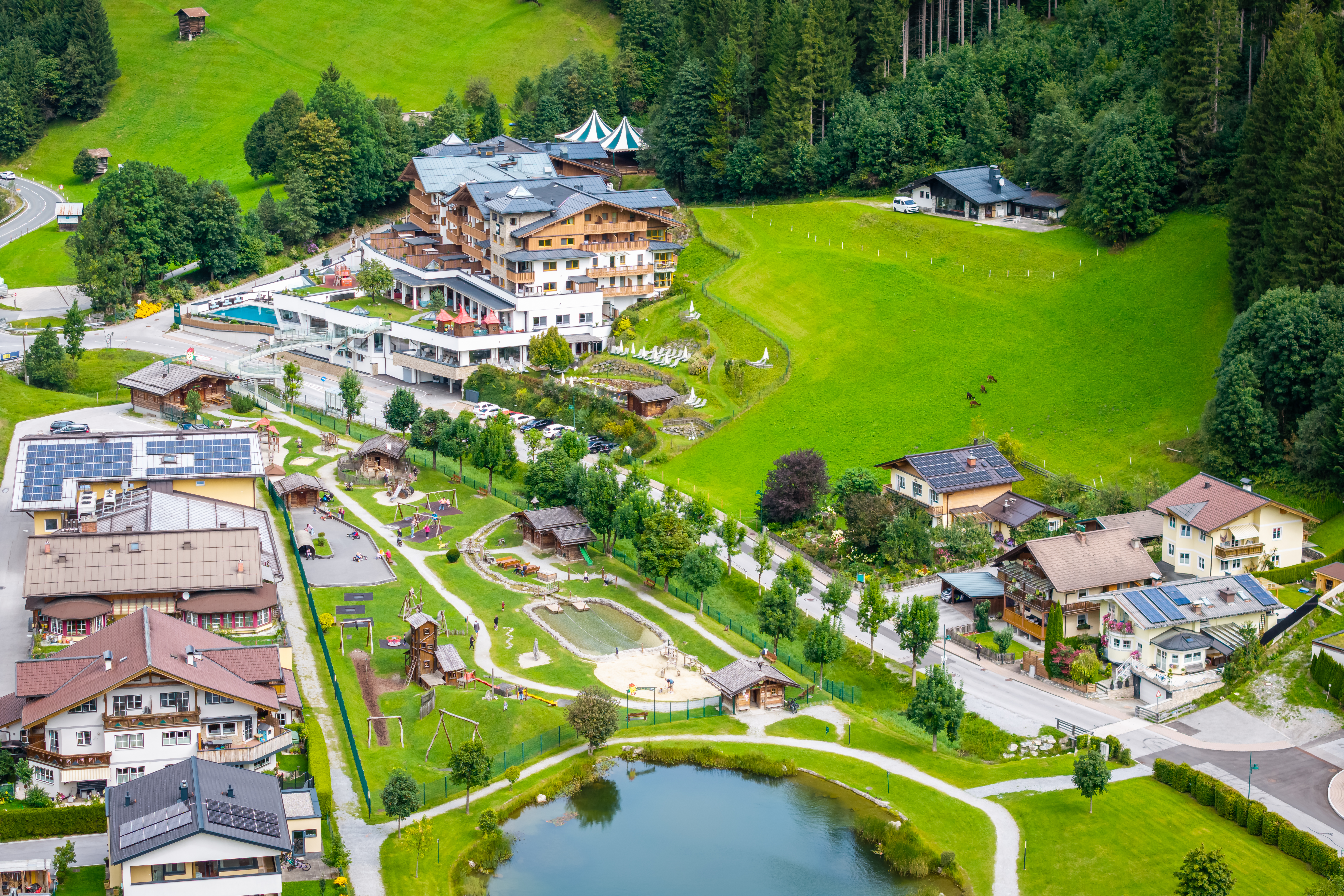 Elevated view of an alpine village with residential houses, a hotel complex, and well-kept green spaces. In the foreground, there is a small pond and a playground, surrounded by meadows and forested hills.