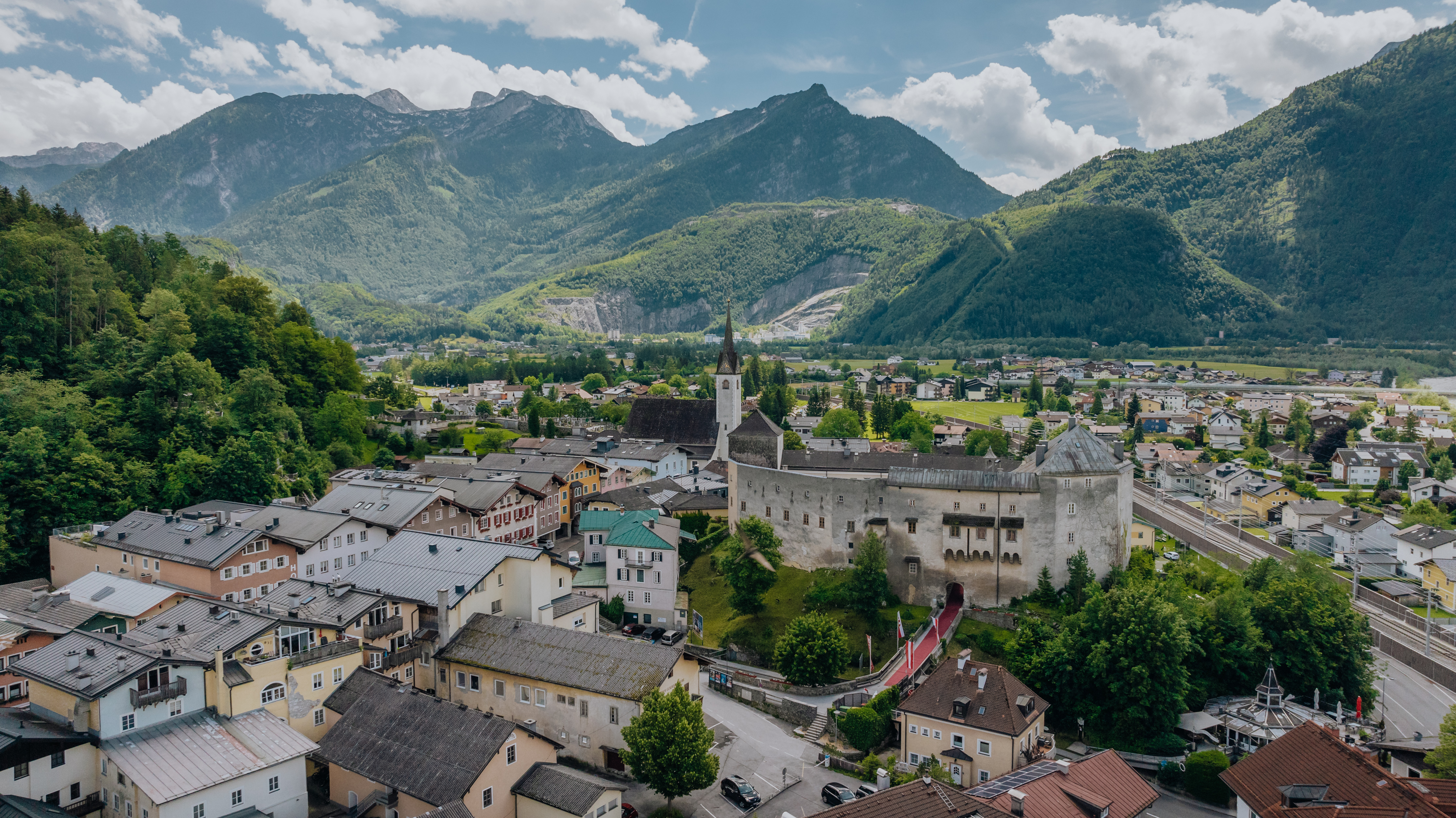 Elevated view of a market town with a historic castle complex and a church steeple at its center. Surrounded by residential houses, a railway line, and green meadows, the town sits against an impressive mountain backdrop under a partly cloudy sky.