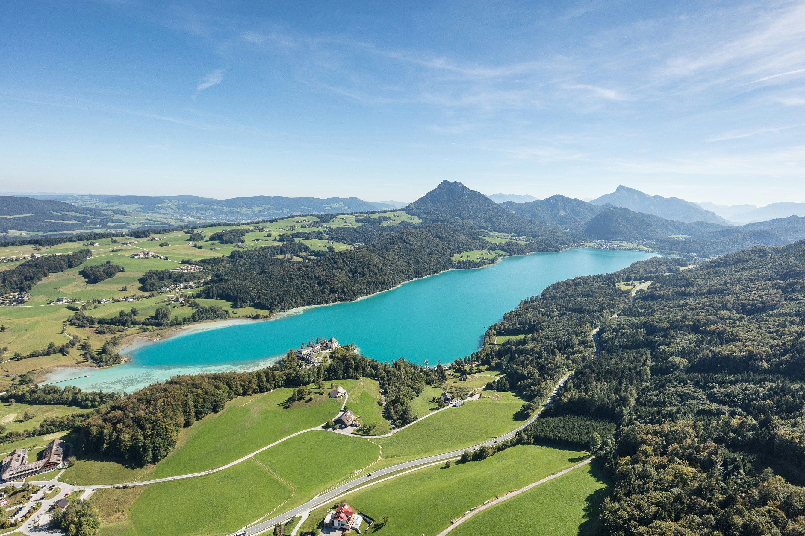 Panoramablick aus der Luft auf einen türkisfarbenen See, umgeben von Wiesen, Wäldern und sanften Hügeln. Am Ufer liegt eine kleine Schlossanlage, dahinter erstrecken sich weite Landschaften und markante Berge unter klarem Himmel.