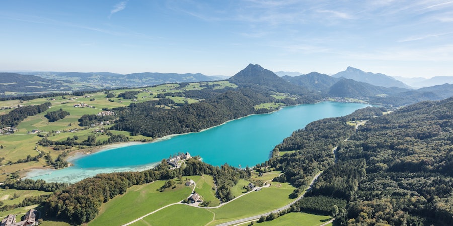 Panoramablick aus der Luft auf einen türkisfarbenen See, umgeben von Wiesen, Wäldern und sanften Hügeln. Am Ufer liegt eine kleine Schlossanlage, dahinter erstrecken sich weite Landschaften und markante Berge unter klarem Himmel.