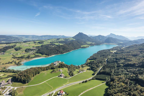 Panoramablick aus der Luft auf einen türkisfarbenen See, umgeben von Wiesen, Wäldern und sanften Hügeln. Am Ufer liegt eine kleine Schlossanlage, dahinter erstrecken sich weite Landschaften und markante Berge unter klarem Himmel.