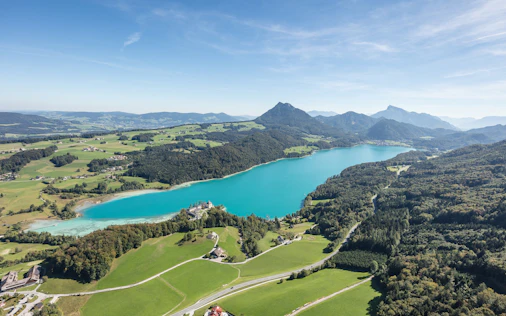 Panoramablick aus der Luft auf einen türkisfarbenen See, umgeben von Wiesen, Wäldern und sanften Hügeln. Am Ufer liegt eine kleine Schlossanlage, dahinter erstrecken sich weite Landschaften und markante Berge unter klarem Himmel.
