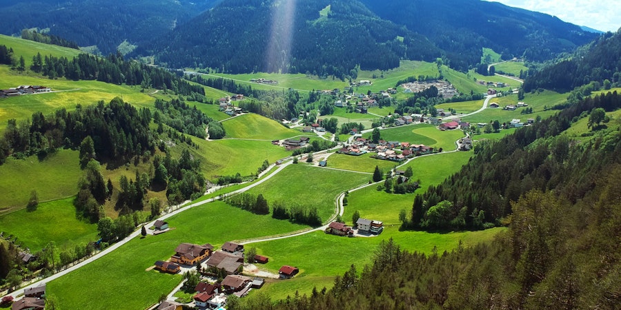 Erhöhter Blick auf ein idyllisches Bergtal mit verstreuten Häusern, grünen Wiesen und bewaldeten Hängen. Kleine Straßen schlängeln sich durch die Landschaft, während im Hintergrund sanfte Berge unter hellem Sonnenlicht aufragen.