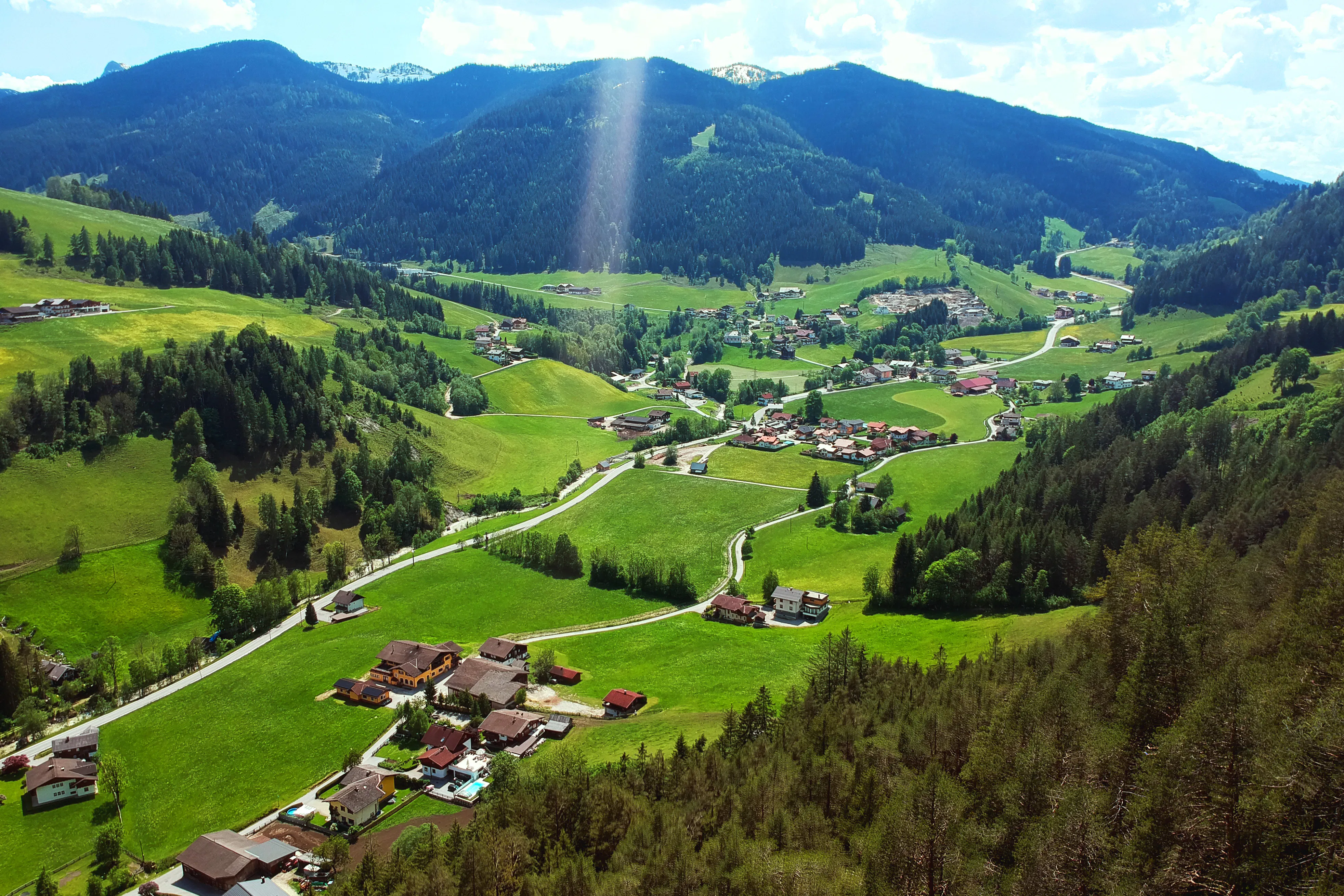 Elevated view of a picturesque mountain valley with scattered houses, green meadows, and forested slopes. Small roads wind through the landscape, while gentle mountains rise in the background under bright sunlight.
